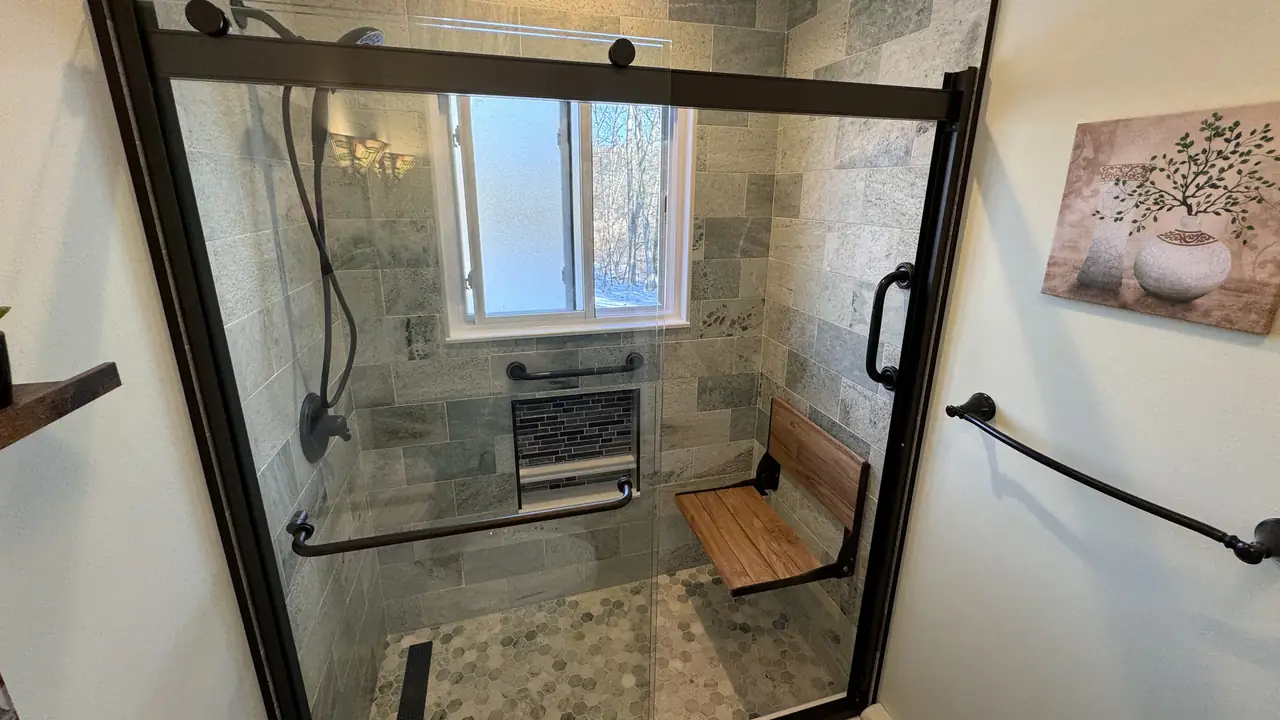 A tiled walk-in shower with a teak fold-down seat, a black grab bar and a mosaic niche